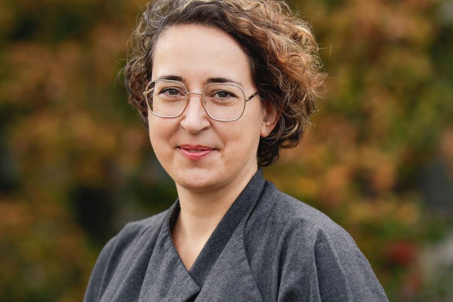Person mit kurzen, lockigen Haaren und grauem Oberteil vor unscharfem Herbsthintergrund.